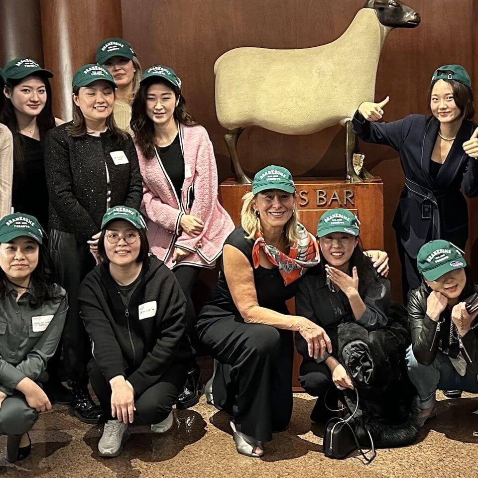 A group of women wearing green baseball caps pose and smile in front of a sheep sculpture inside a building. Most are standing, with a few kneeling in front, and the setting appears to be indoors near a bar area.