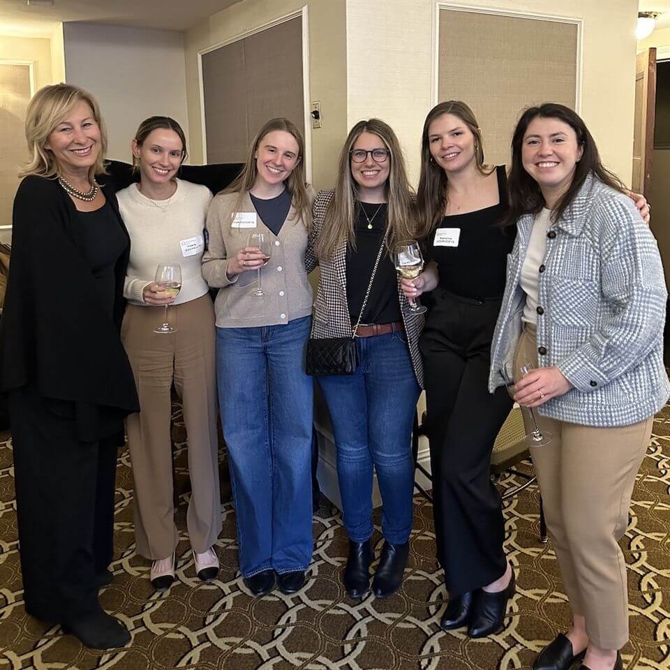 Six women stand together indoors, smiling at the camera. Five of them hold wine glasses. They wear a mix of business-casual outfits and name tags, with a patterned carpet and beige walls in the background.