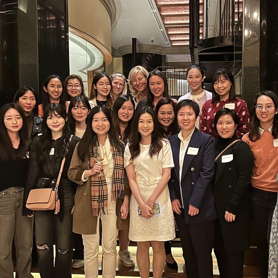 A group of women, dressed in business casual attire, stand closely together and smile for a group photo indoors in front of a staircase and dark glossy walls.