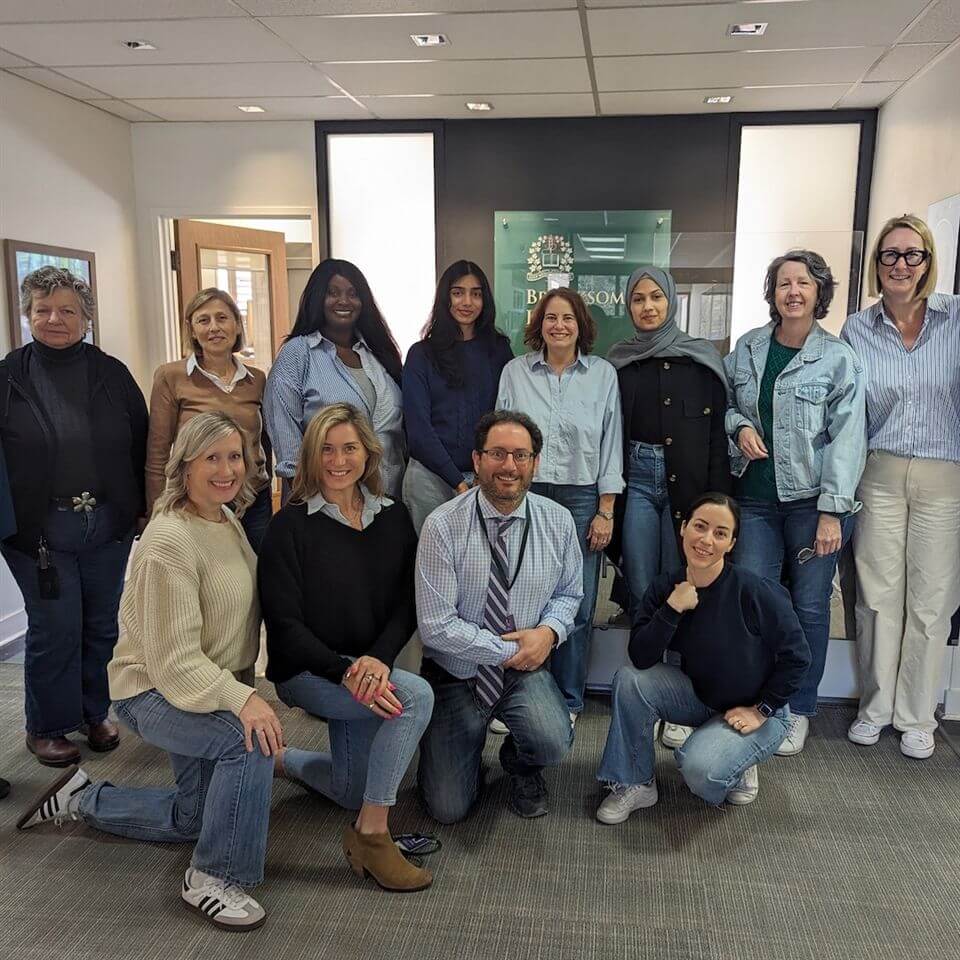 A group of twelve adults, men and women, pose and smile together in an office setting. Most are dressed in casual attire, including blue jeans and light-colored tops. Some are standing, some kneeling, and the mood is friendly.