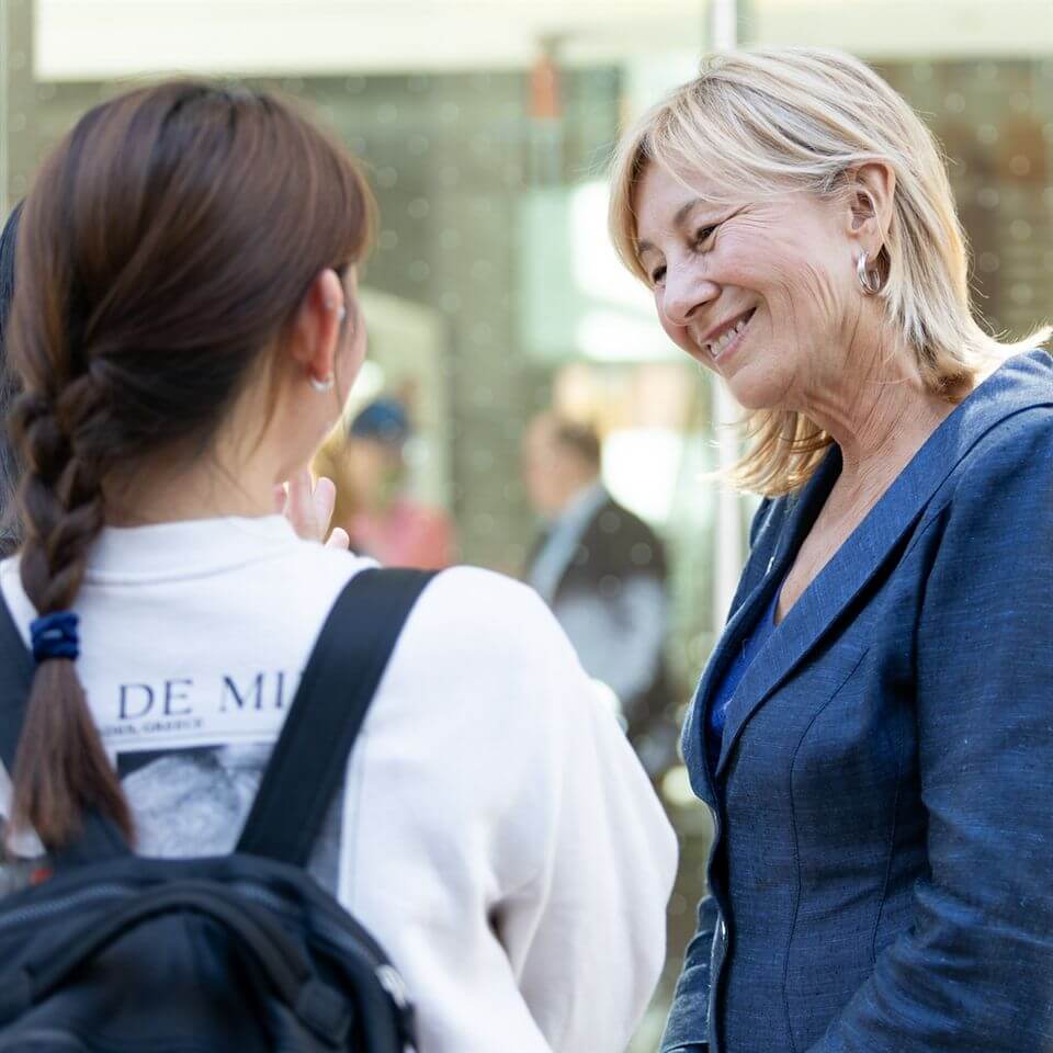 A smiling older woman in a blue blazer talks to a younger person with a braid and a backpack. They appear to be having a friendly conversation outdoors.
