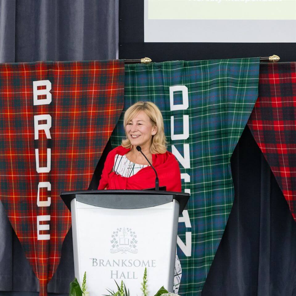 A woman stands at a podium labeled "Branksome Hall," smiling while speaking. Behind her hang two tartan banners with the words "Bruce" and "Duncan" on them.