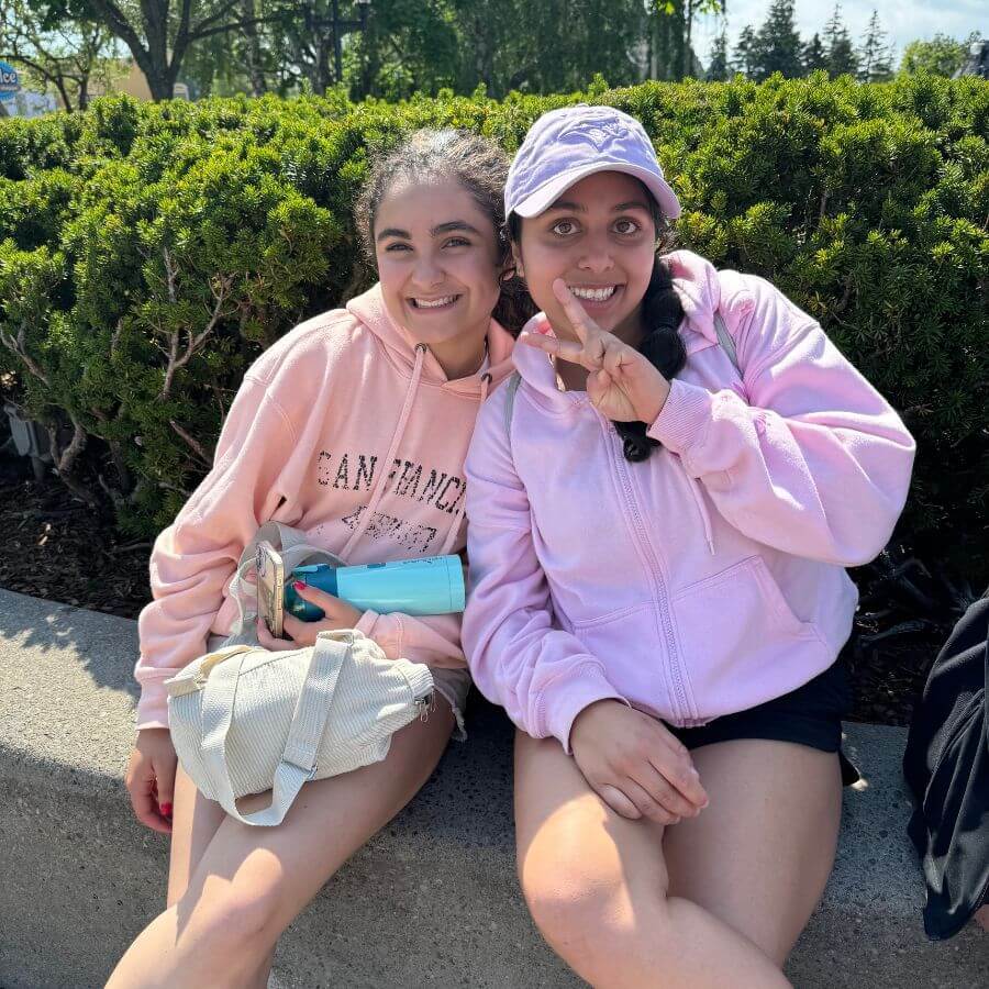 Two young women sit outdoors on a concrete ledge in front of bushes, smiling at the camera. Both wear light pink hoodies; one holds a water bottle and the other flashes a peace sign while wearing a purple cap.