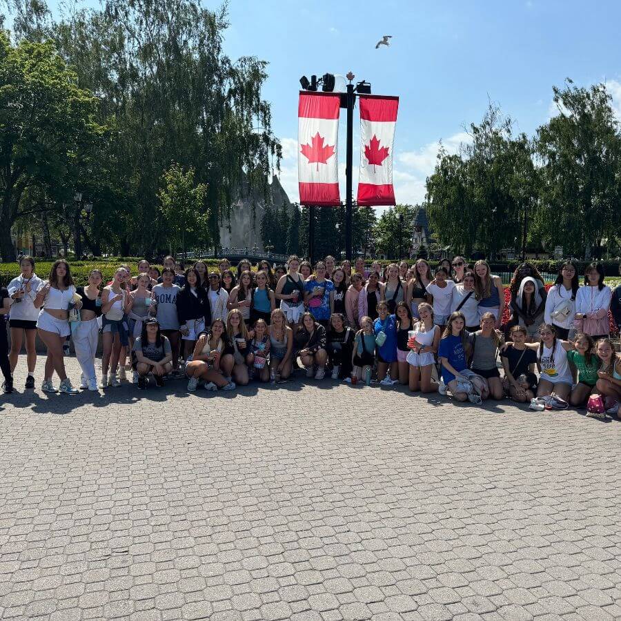 A large group of people poses for a photo outdoors under sunny skies, standing on a paved area with two large Canadian flags on a pole behind them, surrounded by green trees and a clear blue sky.