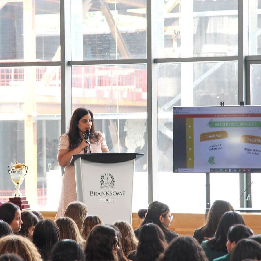 A woman speaks at a podium labeled "Branksome Hall" in front of seated students. A trophy is on a table nearby, and a large screen displays information about athletics representatives. Large windows show an industrial background.