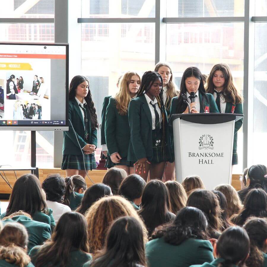 A group of six students in green blazers stand at a podium labeled "Branksome Hall," presenting to an audience. A screen displaying images is visible beside them in a bright room with large windows.