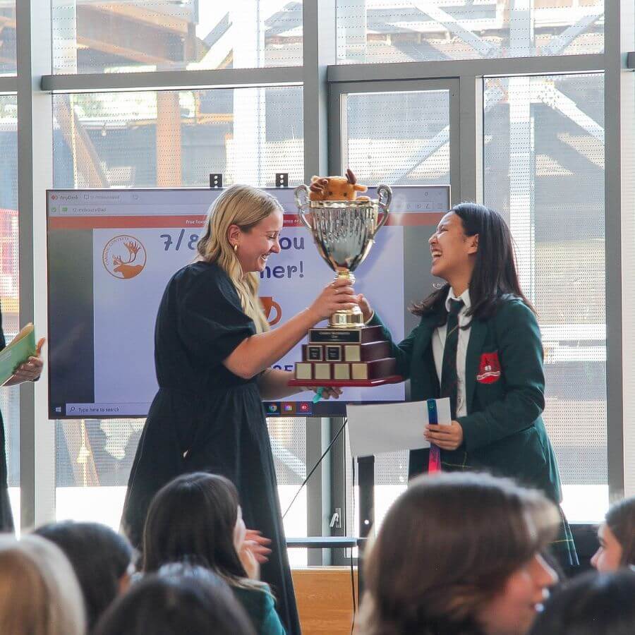 Two women smile and hold a large trophy together during an award ceremony, with an audience watching. A presentation screen in the background displays a congratulatory message.