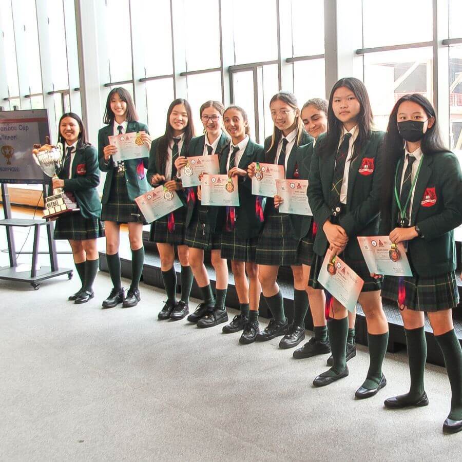 A group of nine students in matching school uniforms stand in a row indoors, holding certificates and medals, smiling at the camera. A trophy is displayed on a table beside them.