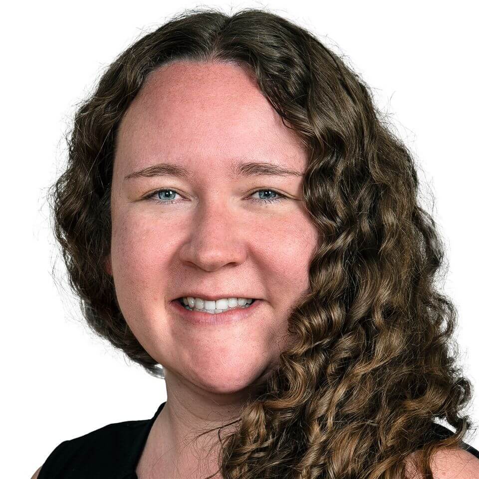 A woman with long, curly brown hair and fair skin smiles at the camera. She is wearing a black top and is posed against a plain white background.