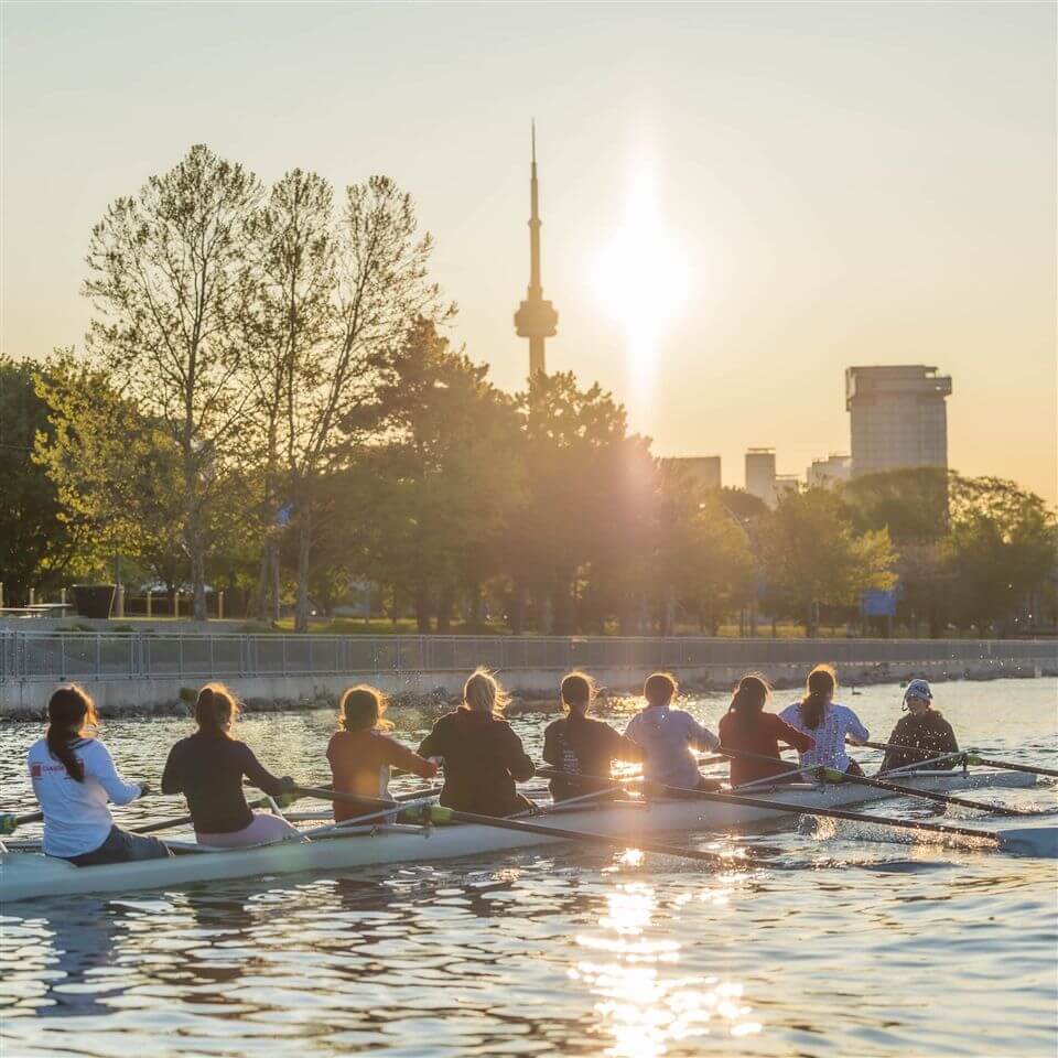 A rowing team paddles on a calm river at sunset, with the city skyline and the CN Tower in the background. Sunlight reflects on the water, and trees line the riverside.