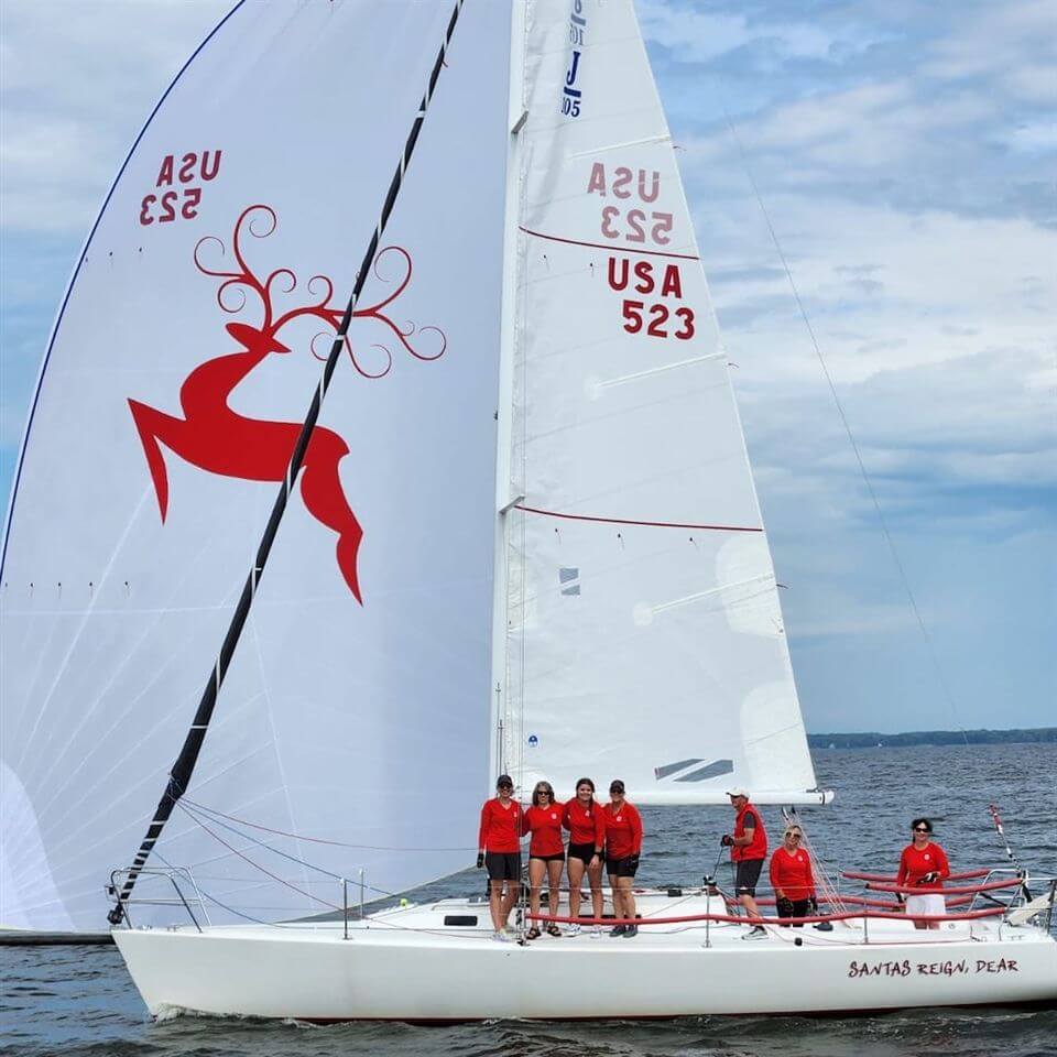 A group of seven people in red shirts stand on a white sailboat with the number 523 and a large red reindeer graphic on the sail, sailing on open water under a partly cloudy sky.