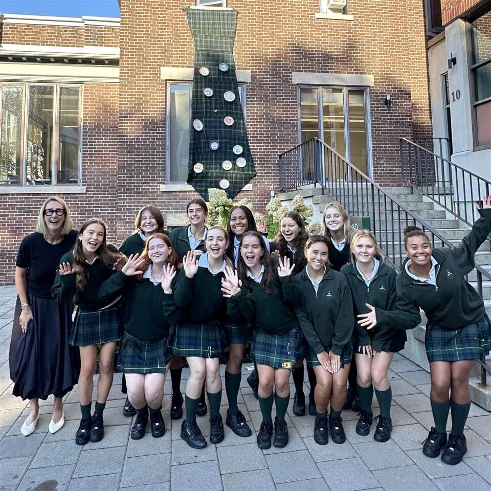 A group of smiling students in green uniforms and a woman in a black skirt stand outside a brick building, posing and waving for the photo. A large decorated tree shape is displayed on the building behind them.