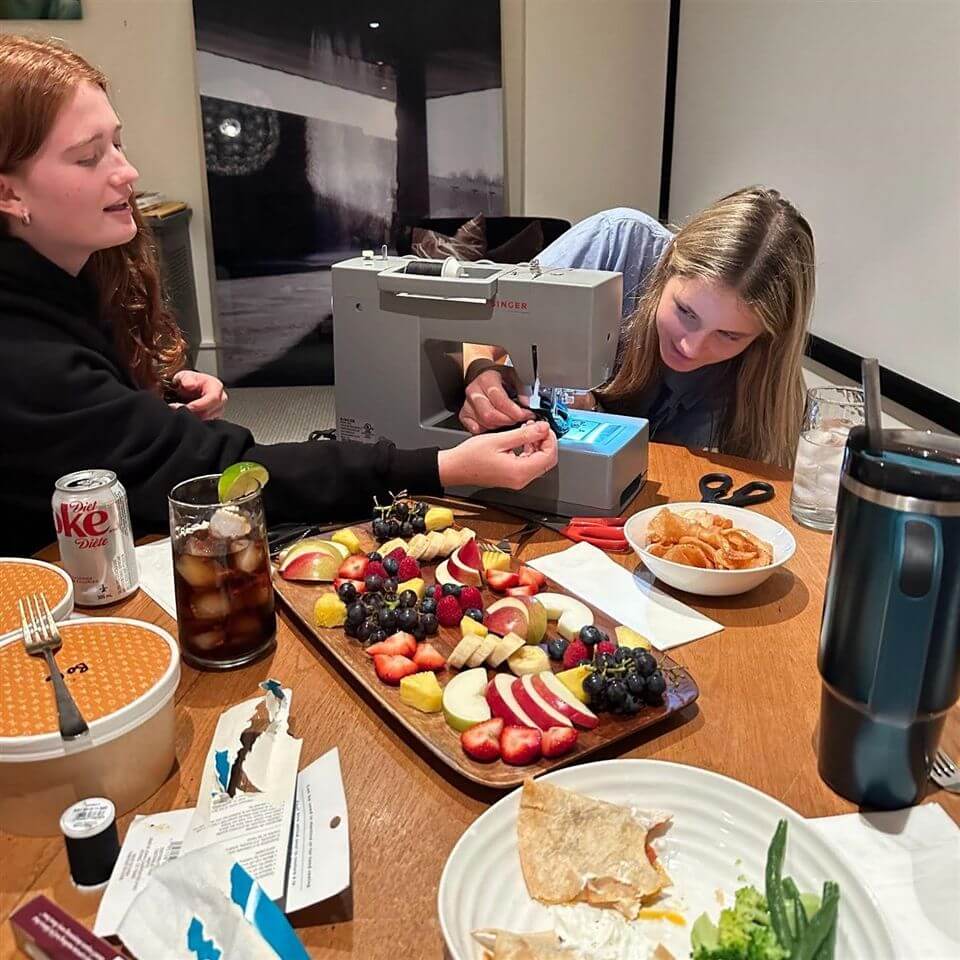 Two young women use a sewing machine at a table covered with snacks, drinks, a fruit and cheese platter, and sewing supplies. One woman focuses on the machine while the other watches closely.