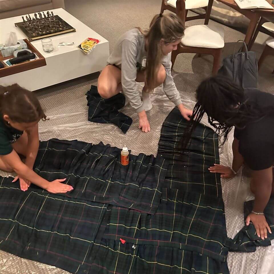 Three people are kneeling on the floor, arranging and gluing pieces of dark plaid fabric together. They are working in a living room with chairs, a table, and a chessboard visible in the background.