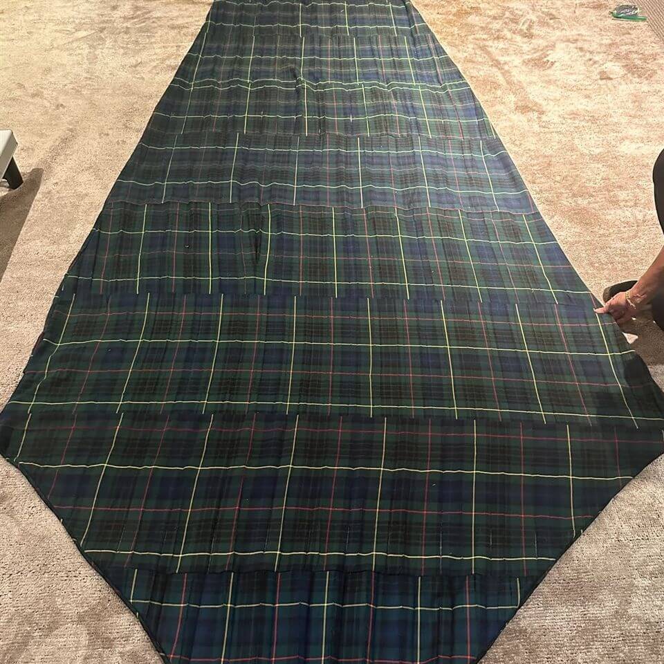 A large piece of dark plaid fabric with green, blue, and red lines is spread out on a carpeted floor. A person’s hand is visible holding one corner of the fabric.