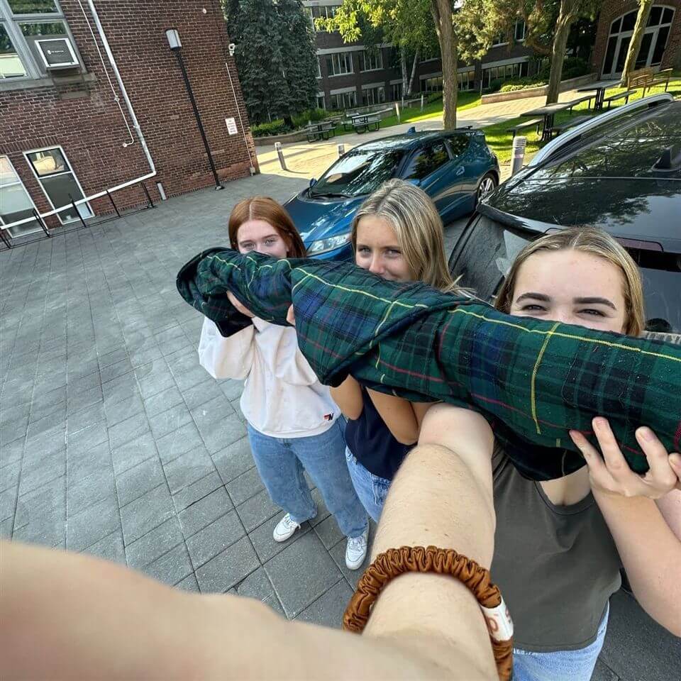 Three young women stand outdoors, smiling and holding a long, plaid-wrapped object between them. They appear to be taking a selfie, with cars and buildings visible in the background.
