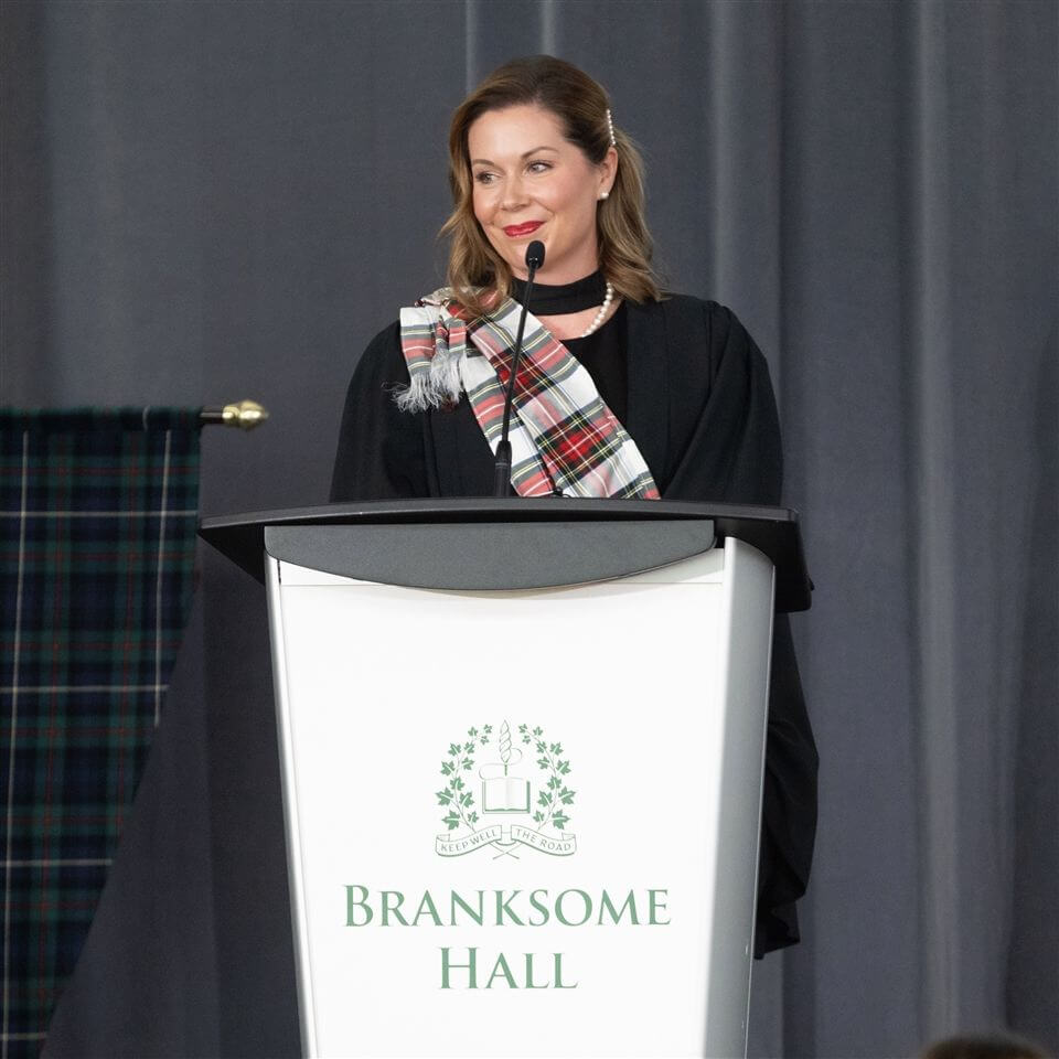 A woman in academic robes and a tartan sash stands at a podium labeled "Branksome Hall," speaking with a smile against a gray curtain backdrop.