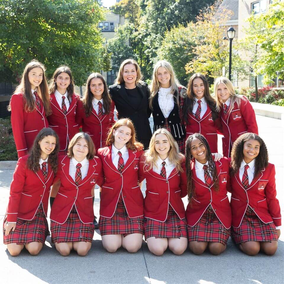 A group of students in red blazers and plaid skirts pose outdoors with two adults, all smiling. Trees and a building are visible in the background, suggesting a school setting.