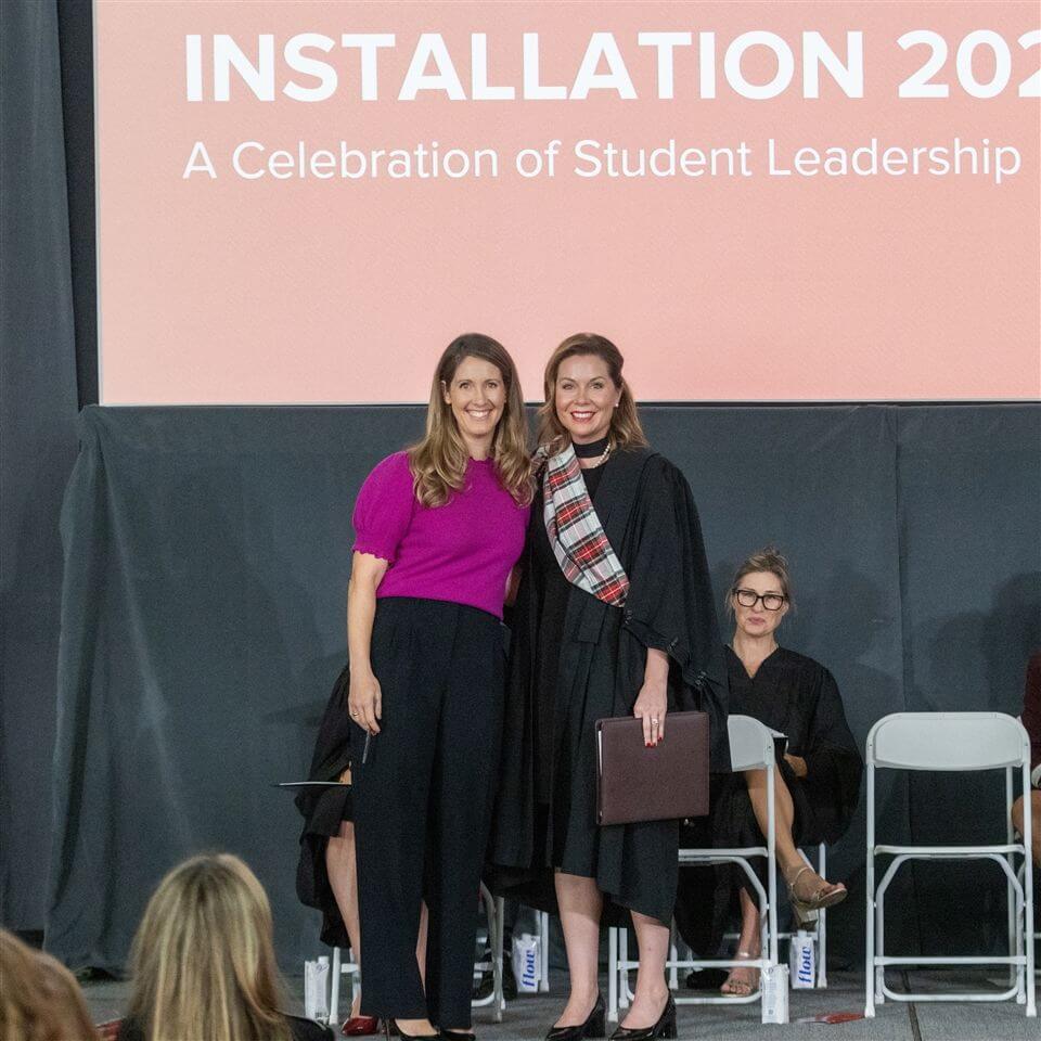 Two women stand and smile in front of a stage backdrop reading "Installation 2023: A Celebration of Student Leadership." One wears black academic robes and a plaid sash, while the other wears a magenta top and black pants.