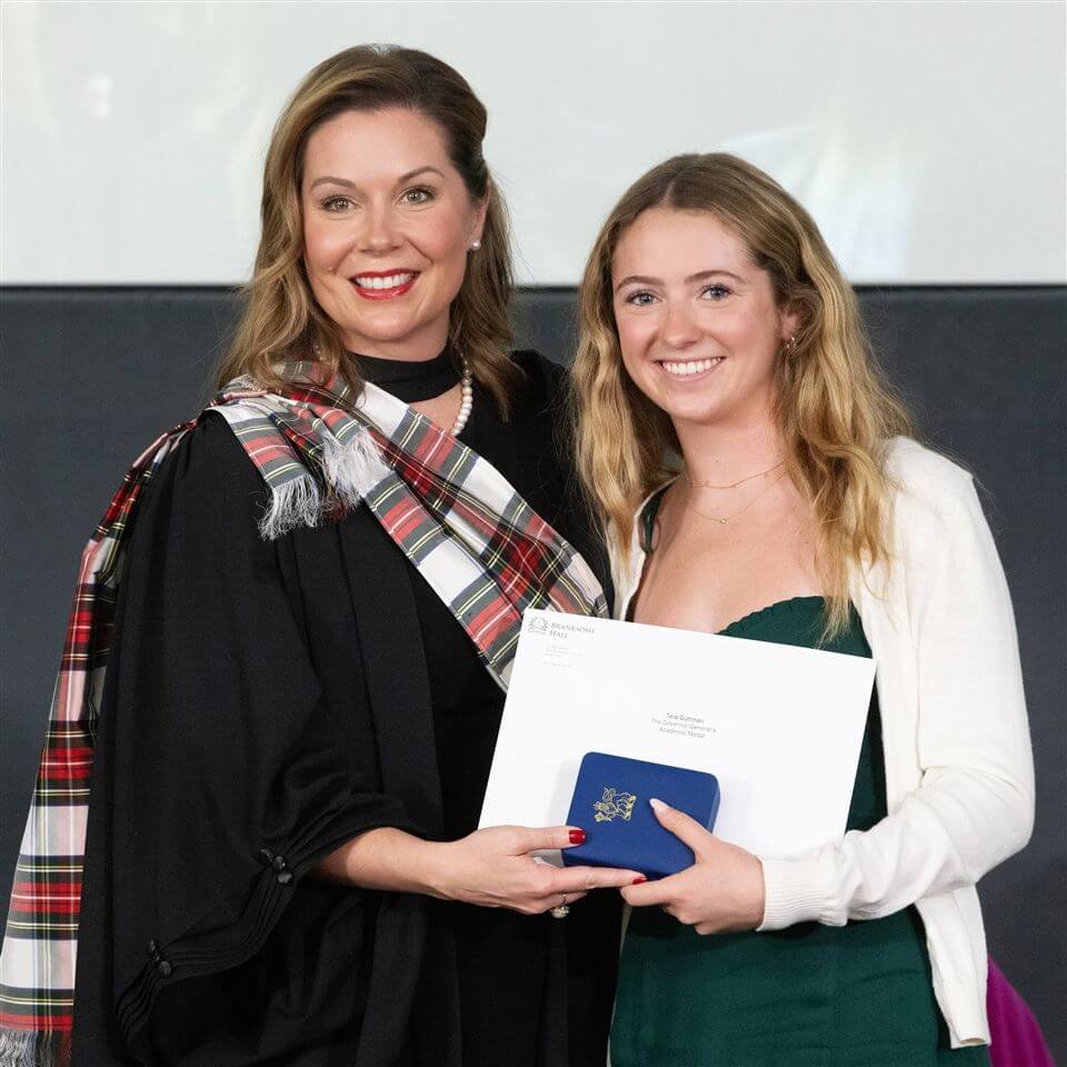 Two women smiling at a ceremony, one holding a certificate and a blue box. The woman on the left wears a black robe with a tartan sash, while the woman on the right wears a green dress and white sweater.