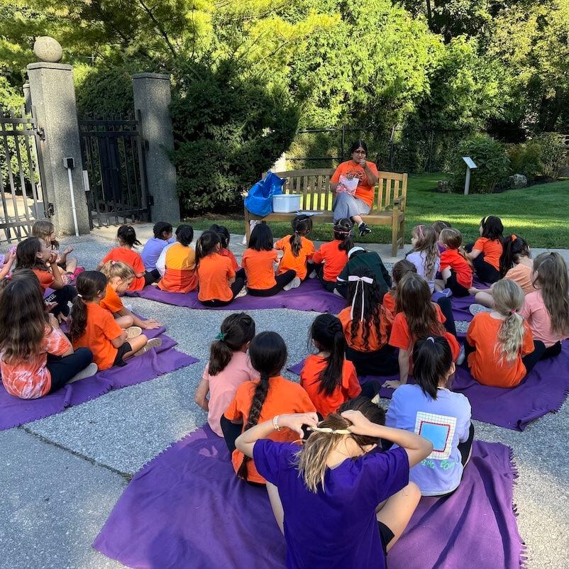 A group of children sit on purple mats outdoors, facing a woman who is speaking to them near a bench. Most of the children wear orange shirts, and the scene is shaded by trees and surrounded by greenery.