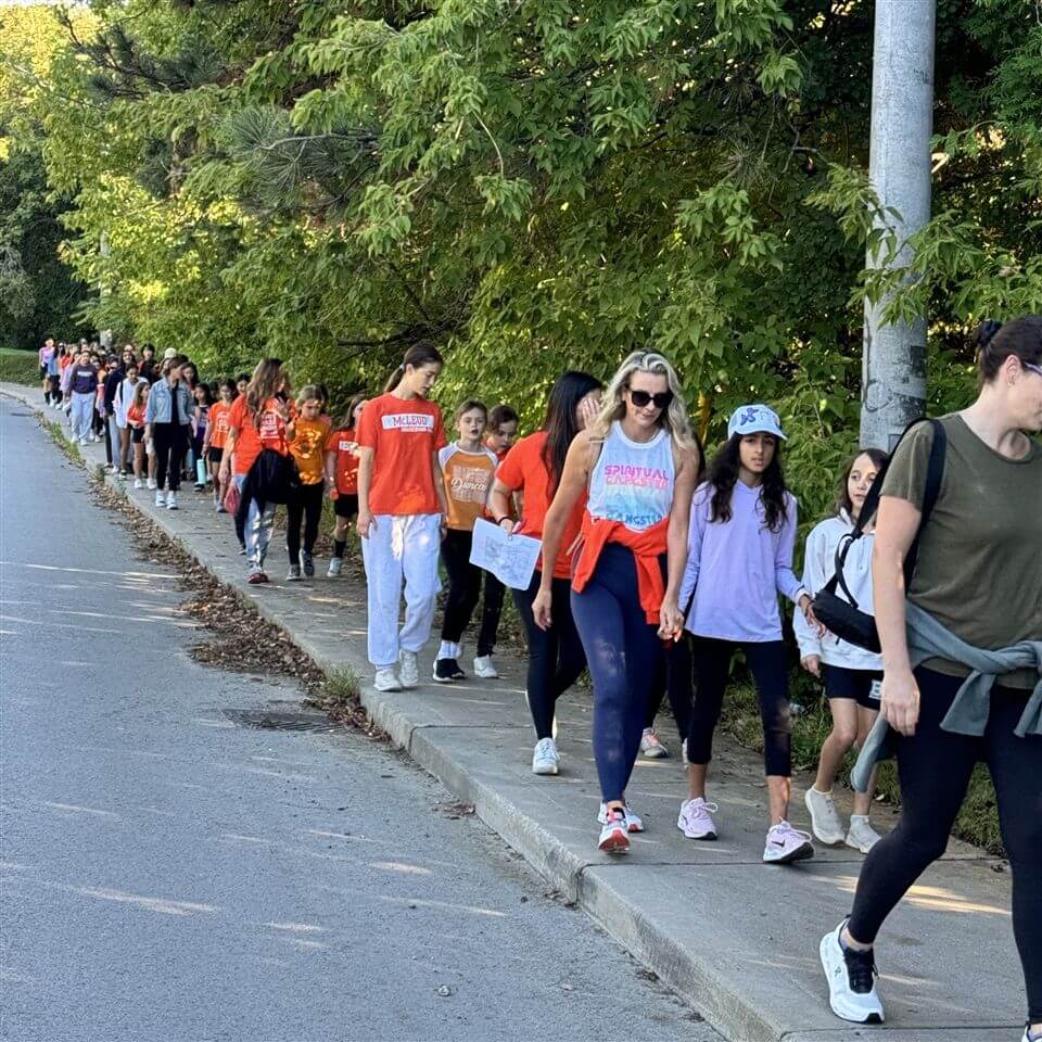 A group of people, including children and adults, walk in a long line along a sidewalk bordered by trees. Many are wearing orange shirts, and the atmosphere appears calm and organized.