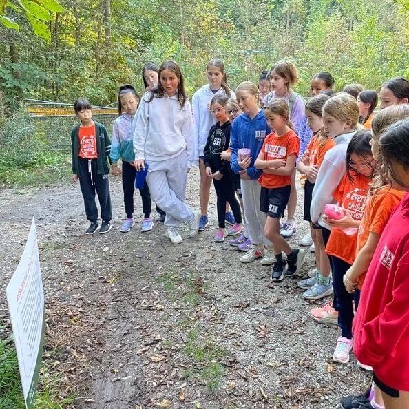 A group of children and a few adults stand outdoors on a wooded path, gathered around a sign with text, appearing engaged and attentive. They are dressed in casual clothing and sneakers.