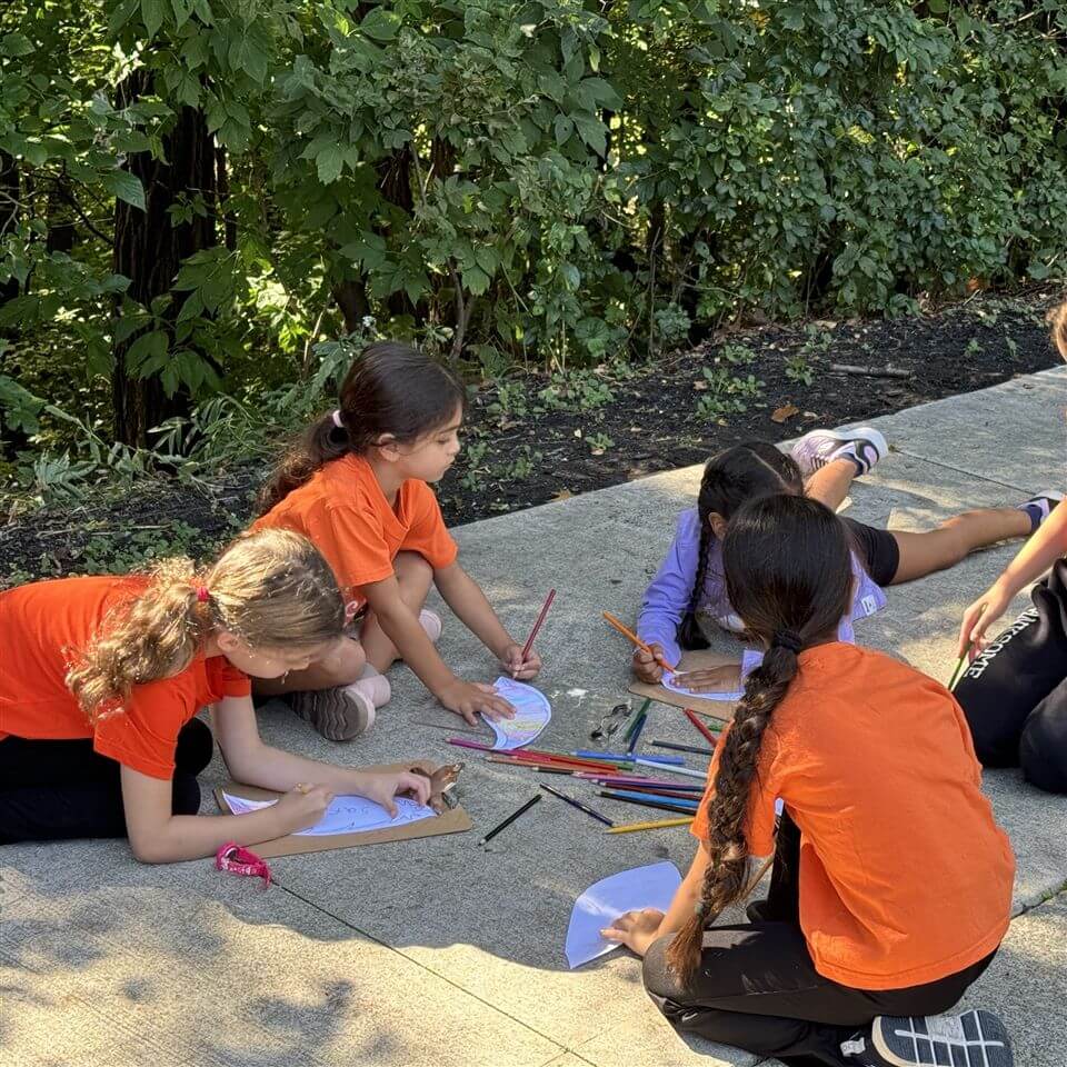 Four young girls sit on a sidewalk outdoors, surrounded by trees, drawing with colored pencils on paper. They wear orange shirts and focus on their art, with supplies scattered around them.