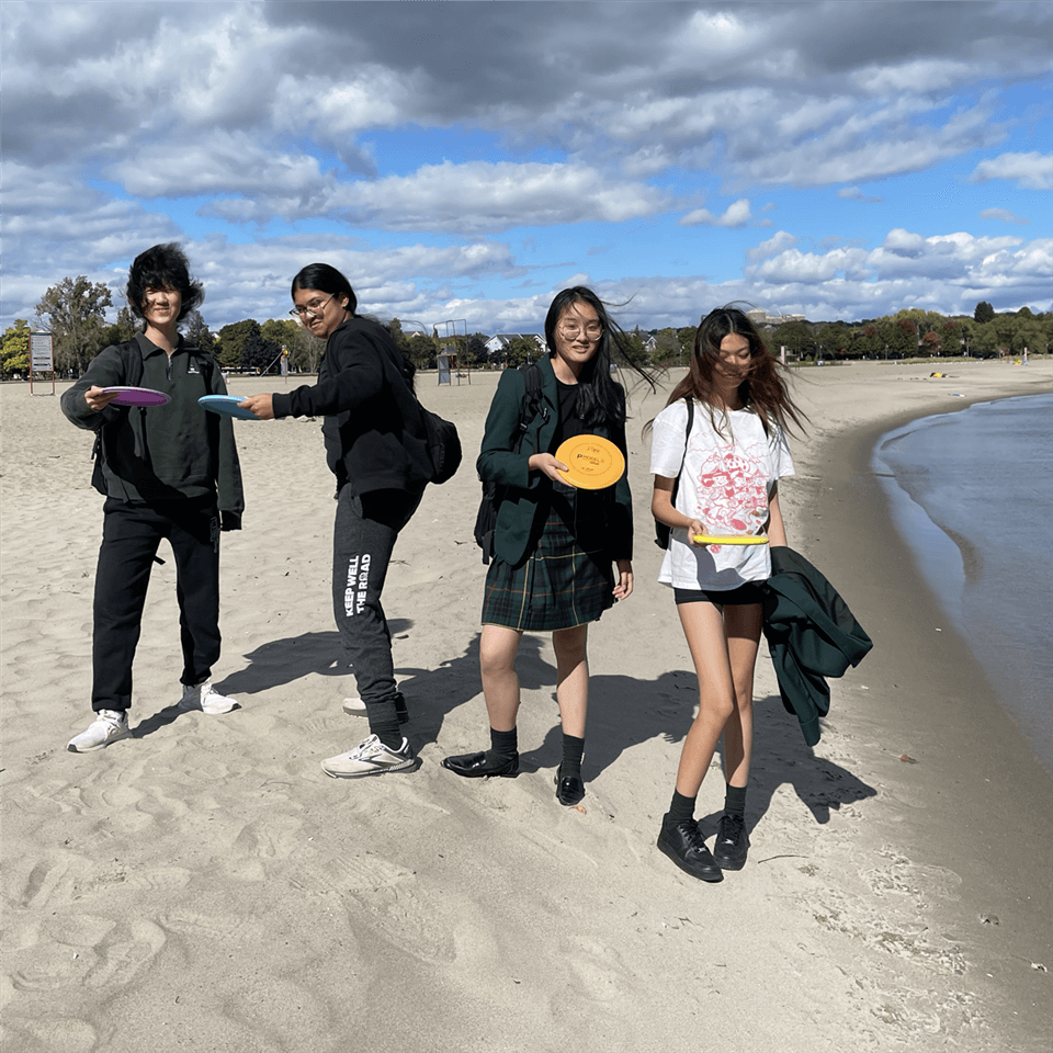 Four young people stand on a sandy beach under a partly cloudy sky, each holding a frisbee and smiling toward the camera. The shoreline and trees are visible in the background.