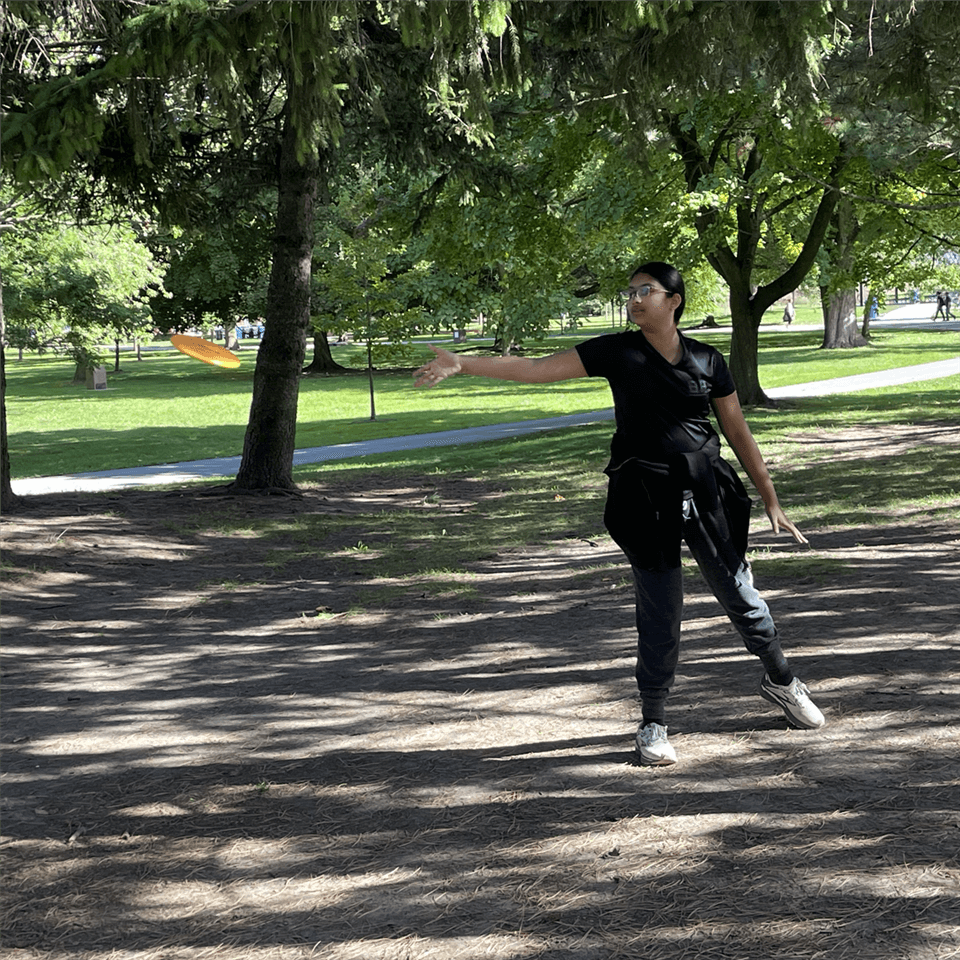 A person wearing black clothes throws an orange frisbee in a park with tall trees and green grass. Sunlight filters through the foliage, casting shadows on the ground.