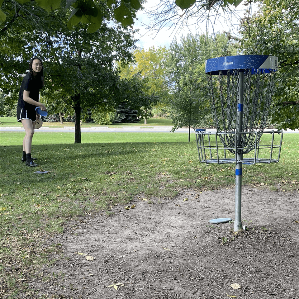 A person stands outdoors on a grassy area, preparing to throw a disc towards a disc golf basket. Trees and a road are in the background under a partly cloudy sky.