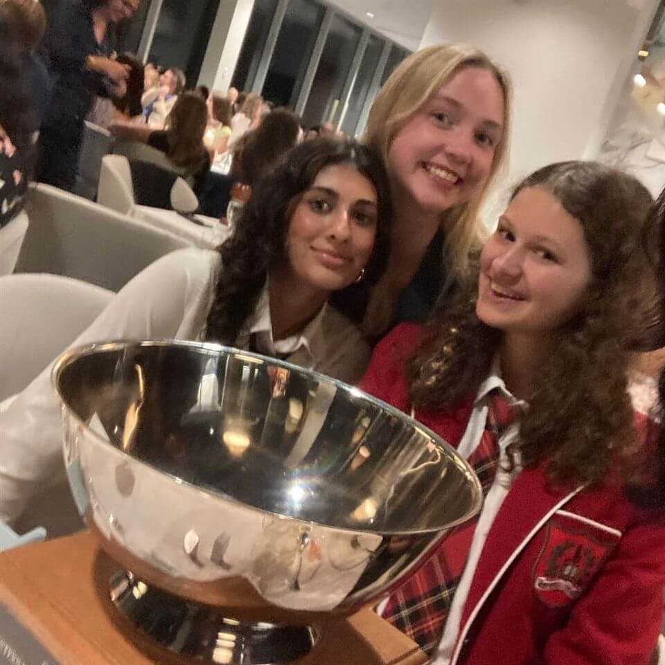 Three smiling young women, two in school uniforms, sit closely together at a table. In front of them is a large silver trophy bowl. The background shows people seated at tables in a bright, modern room.