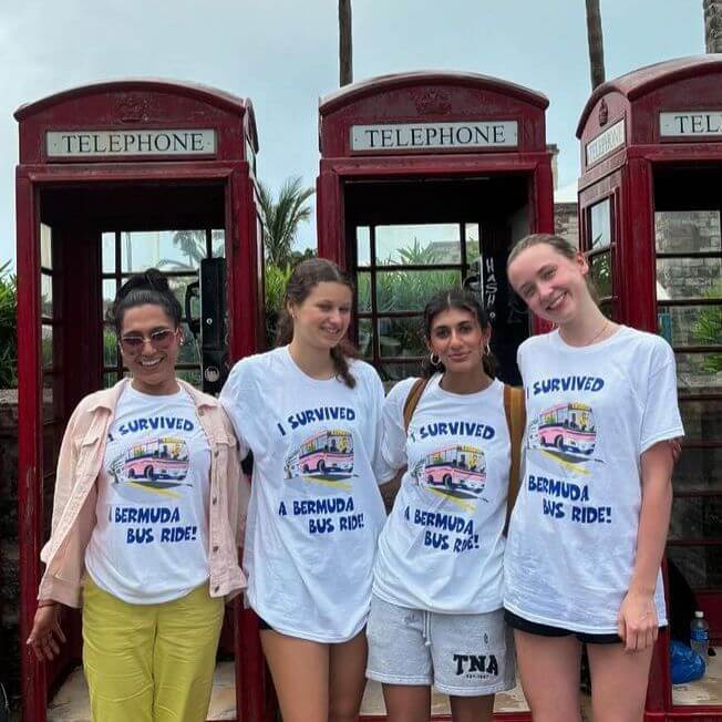 Four smiling women stand in front of three red telephone booths, wearing matching white T-shirts that read "I survived a Bermuda bus ride!" with a colorful bus graphic.