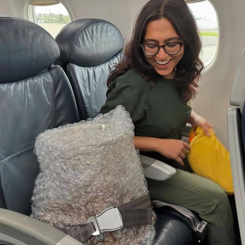 A woman smiling on an airplane, sitting next to a seat with a large bubble-wrapped package secured with a seatbelt.