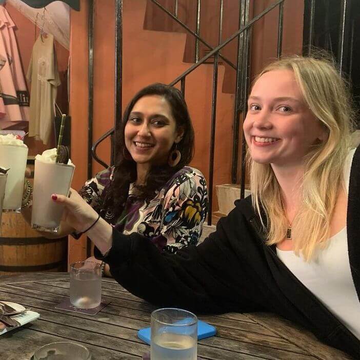 Two women sitting at a wooden table indoors, smiling and holding up milkshakes in celebration. There are glasses of water on the table and a spiral staircase in the background.