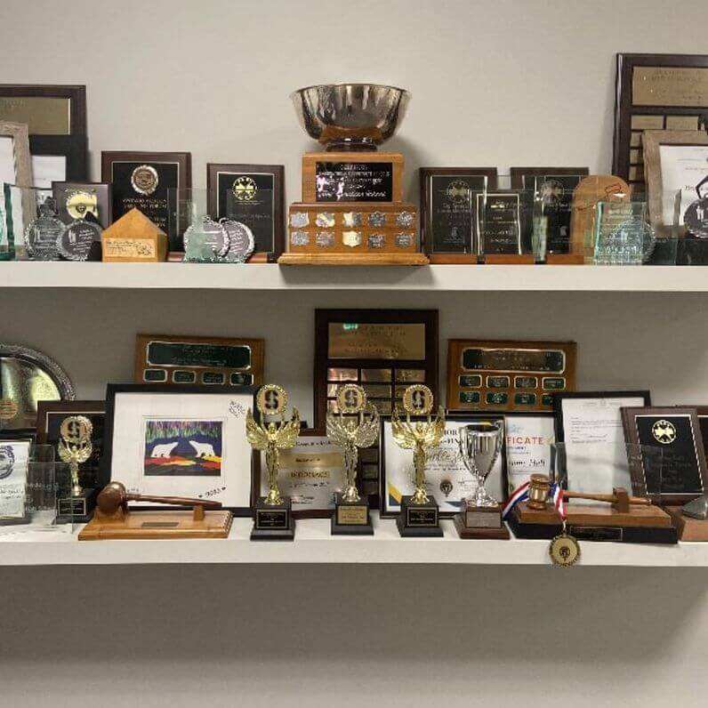 Two white shelves display a collection of trophies, plaques, certificates, and various awards, made mostly of wood, metal, and glass, all neatly arranged against a plain wall.