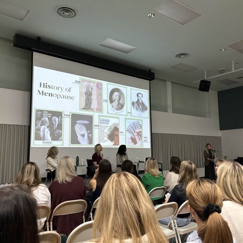 A group of people sit facing a presentation titled "History of Menopause," featuring historical images and text, while a speaker stands at the front and three panelists sit on stage.