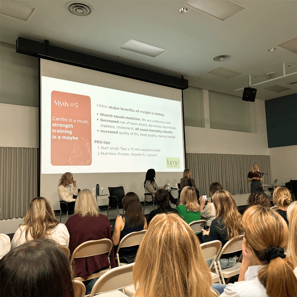 A woman gives a presentation to a seated audience in a classroom. A slide on the screen debunks the myth “Cardio is a must, strength training is a maybe,” and lists benefits and tips for weight training.
