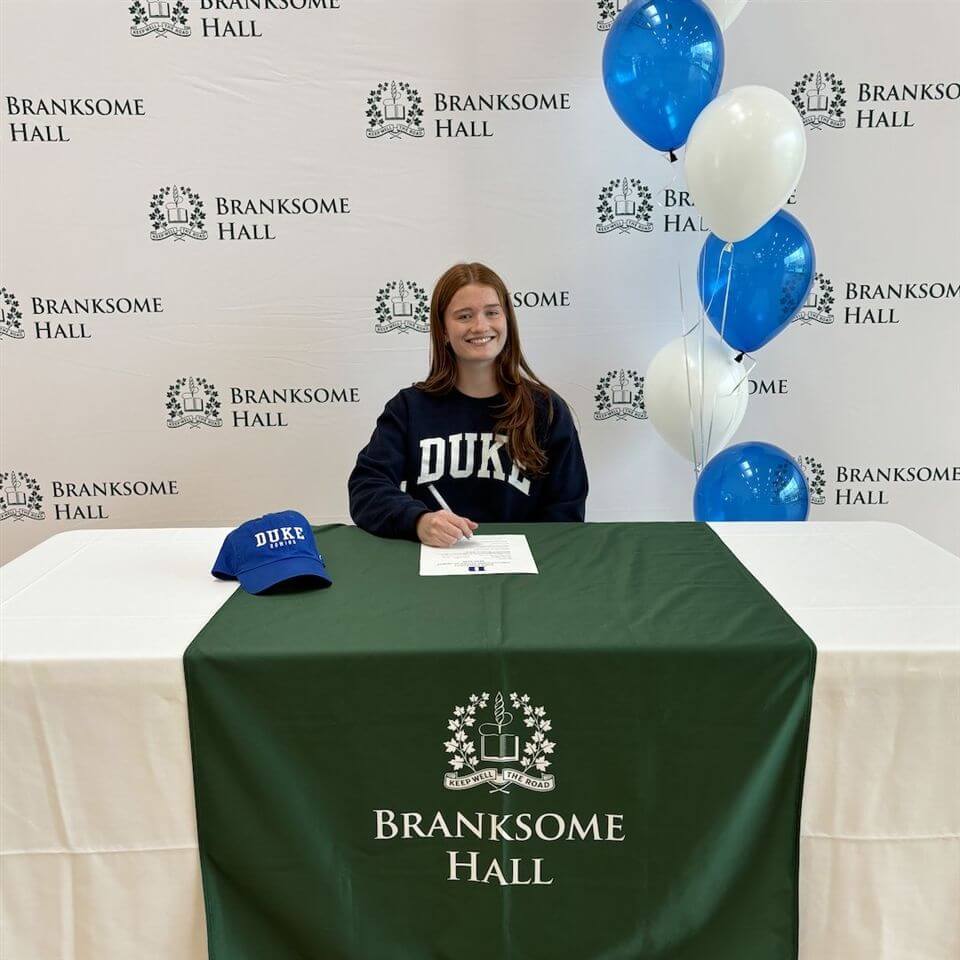 A smiling young woman sits at a table with a "Branksome Hall" cloth, signing a paper. She wears a "Duke" sweatshirt, with a Duke hat and blue and white balloons next to her. Branksome Hall logos are in the background.