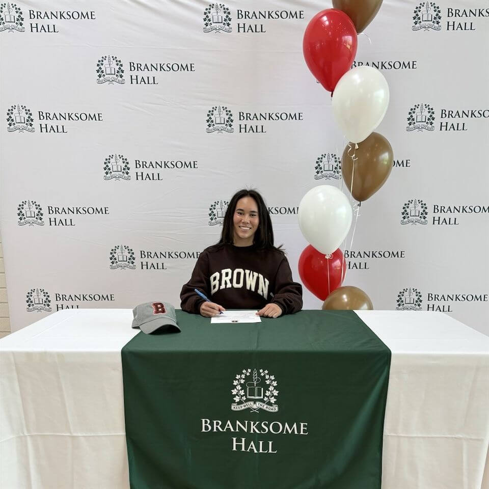 A young woman in a "Brown" sweatshirt sits at a table with a Branksome Hall tablecloth, signing a paper. Red, white, and brown balloons and a gray cap are on the table. A Branksome Hall backdrop is behind her.