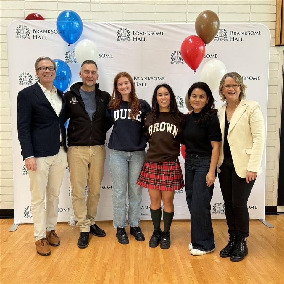 Six people stand smiling in front of a Branksome Hall backdrop with red, blue, and brown balloons; two young women in college shirts (Duke and Brown) are flanked by four adults, all dressed in business casual attire.