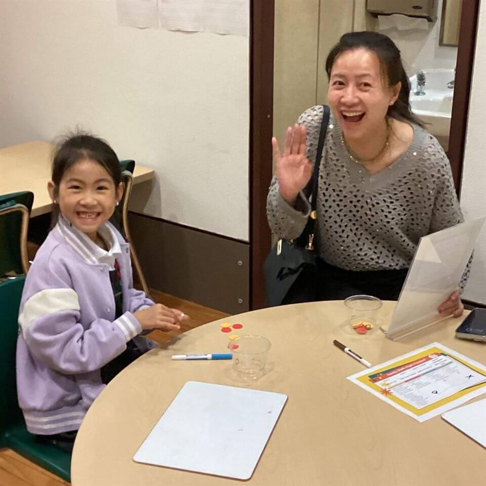 A smiling young girl and a woman wave at the camera while sitting at a round table with game pieces, a whiteboard, and papers, in a classroom or learning environment.