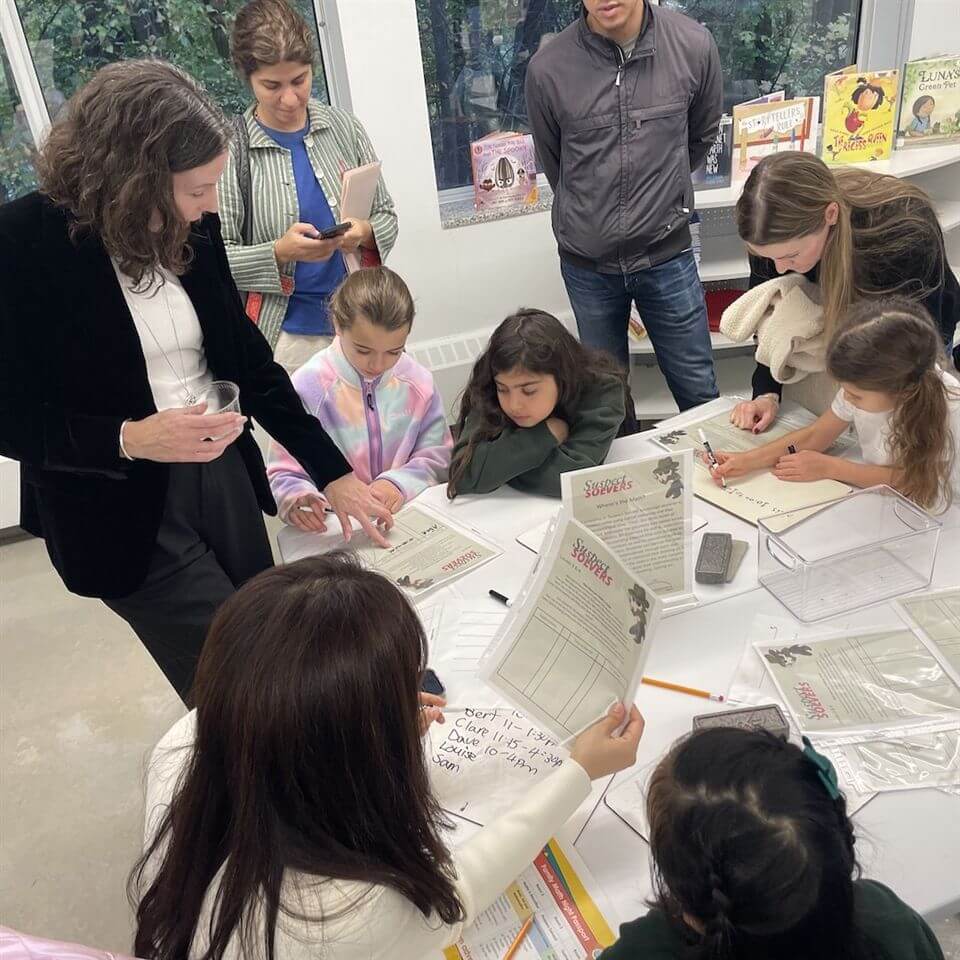 A group of adults and children gather around a table covered with papers and clipboards, engaging in writing or drawing activities in a brightly lit room with books displayed in the background.
