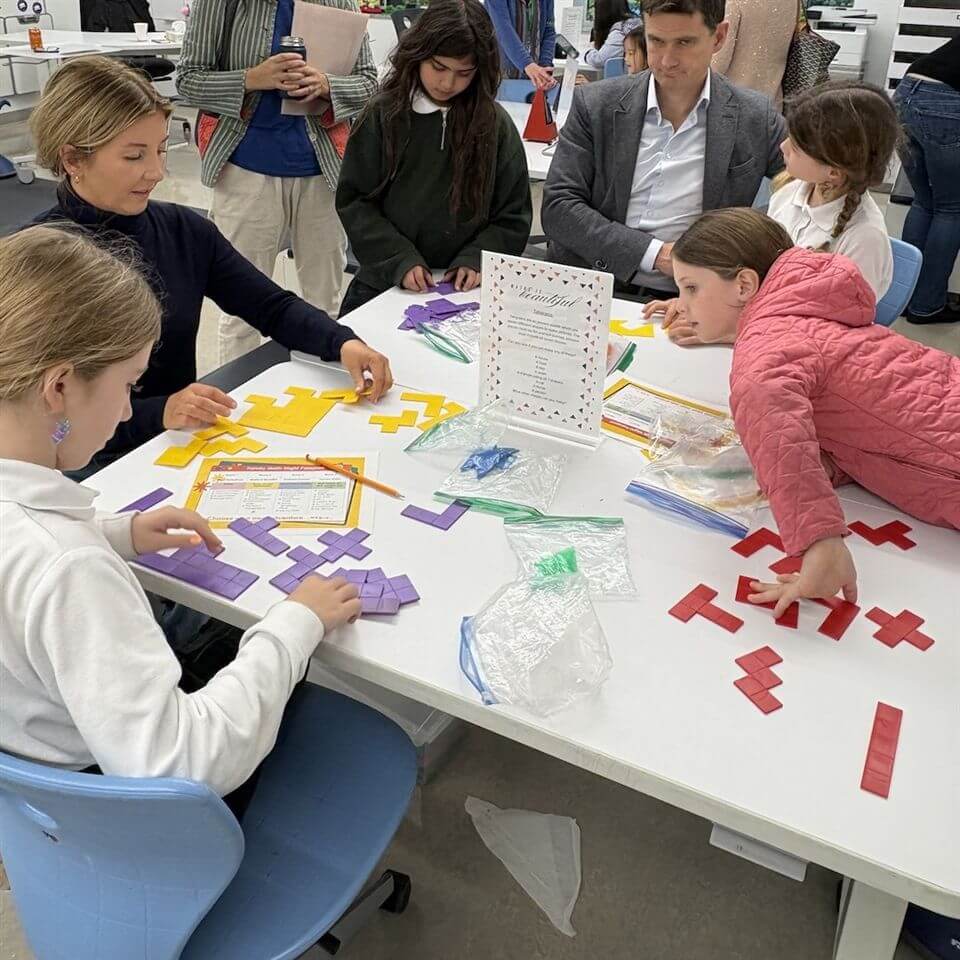 A group of children and adults sit around a table, working with colorful geometric puzzle pieces. Papers and plastic bags with more pieces are on the table, and a sign stands in the center. The environment looks like a classroom.