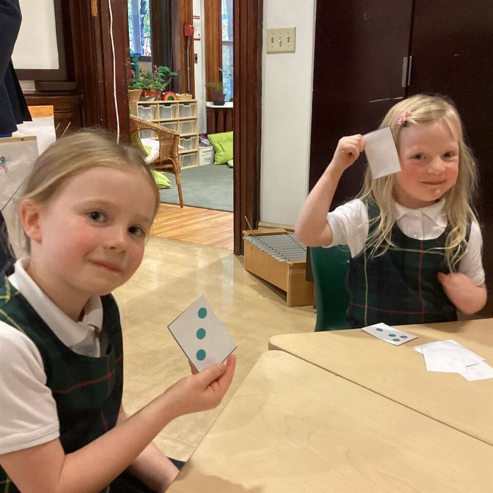 Two young girls in school uniforms sit at a table, each holding up a card with dots on it. One card shows three blue dots. Both girls are smiling inside a bright classroom with wooden furniture and plants.