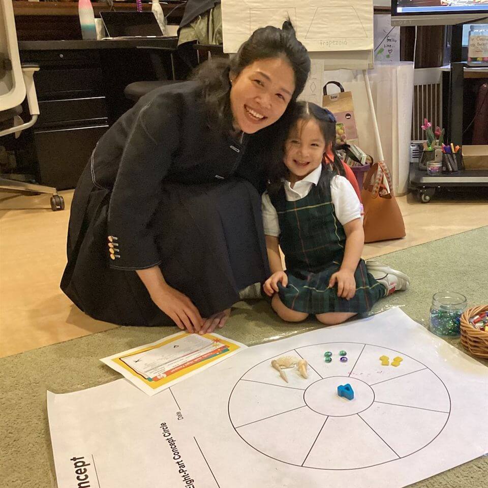 A smiling woman and young girl kneel on the floor next to a large paper game board with colorful game pieces. The girl wears a plaid dress and white shirt; both look happy in a cozy indoor setting.
