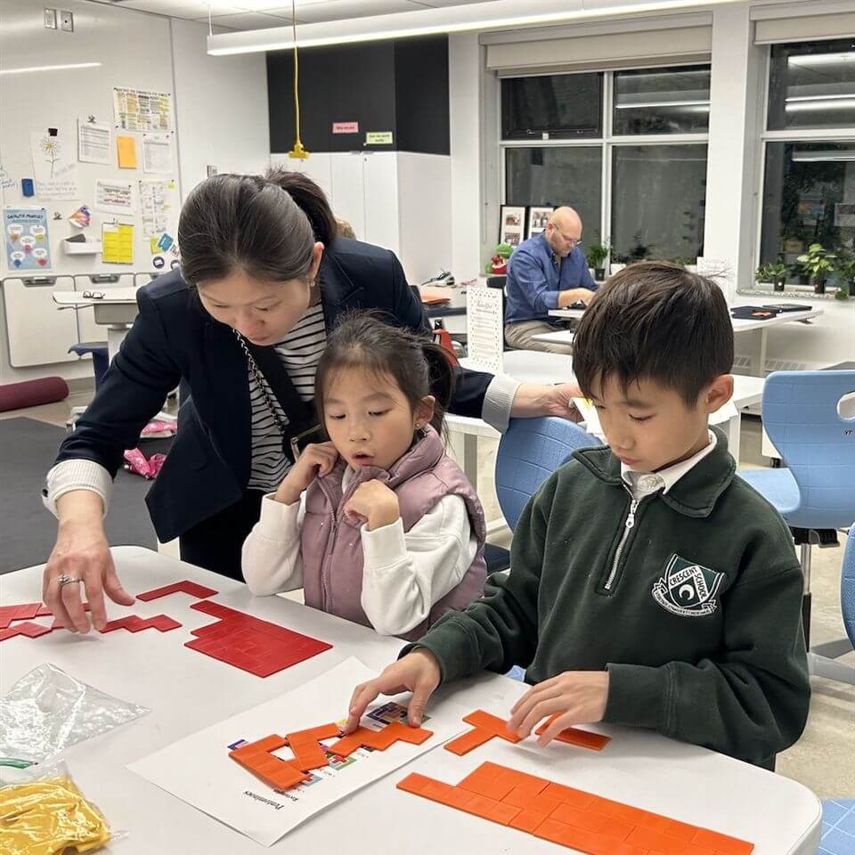 A woman helps two young children arrange red puzzle pieces on a table in a bright classroom, while another adult works in the background. The children look focused on the activity.