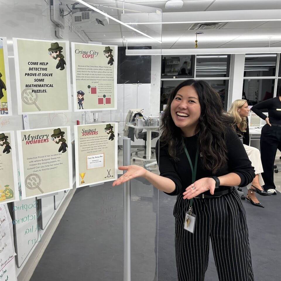 A woman smiles and gestures toward a display board with colorful posters about mathematical puzzles and a detective theme, set in a bright, modern classroom. Another person sits in the background.