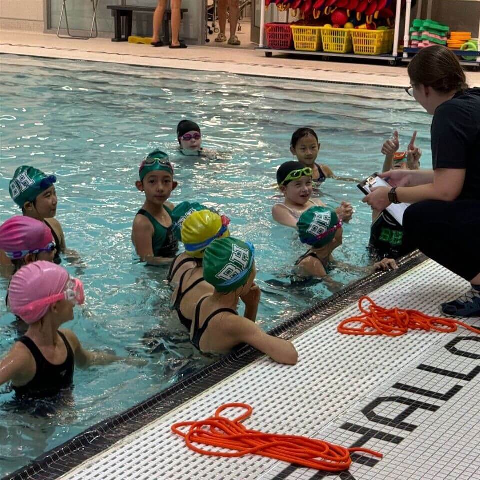 A group of children wearing swim caps and goggles are gathered in a swimming pool next to the edge, listening attentively to an instructor sitting poolside. Orange ropes are coiled on the pool deck.