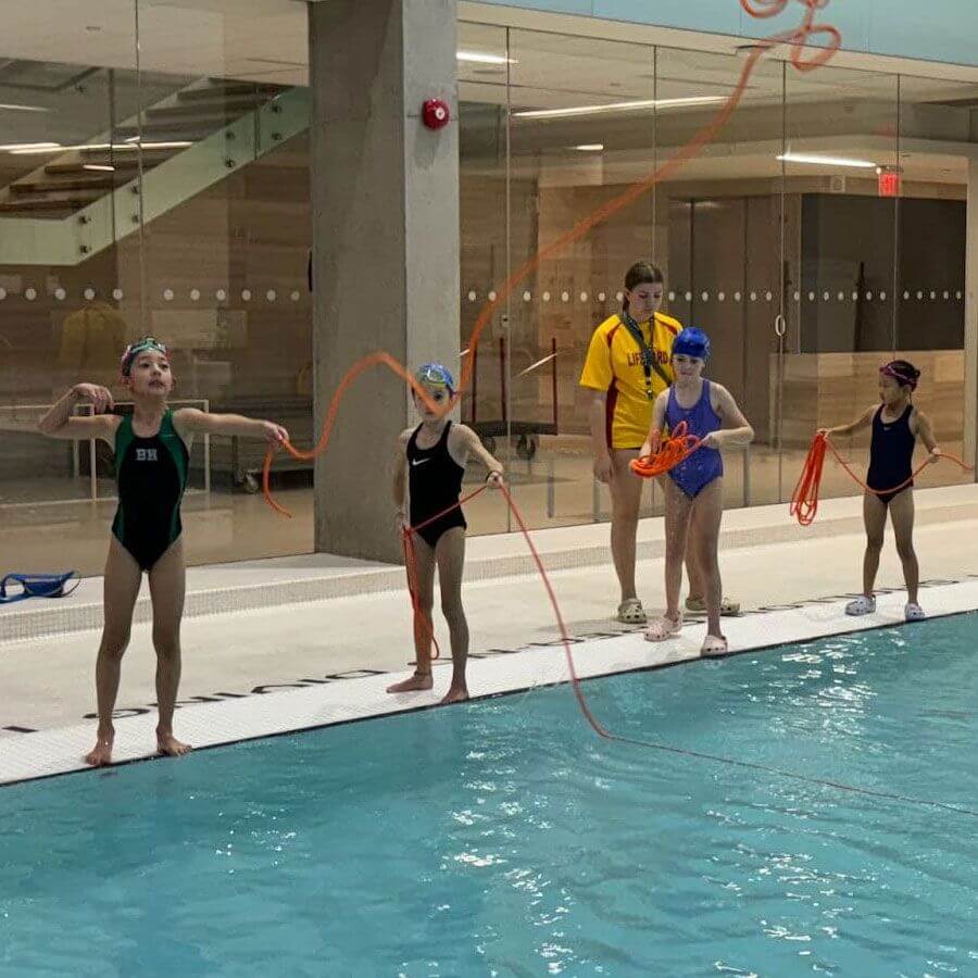 Four young swimmers stand by an indoor pool, throwing orange ropes into the water, supervised by a coach in a yellow shirt. The pool area has glass walls and visible stairs in the background.